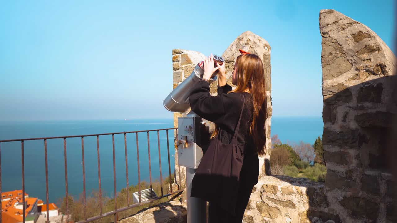 Young woman with long brown hair reaching for binoculars and looking through them on rooftops and the sea