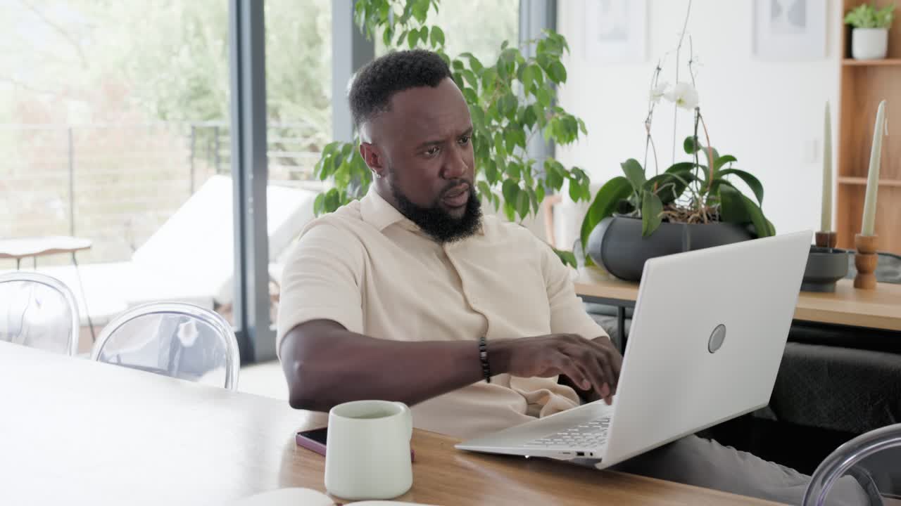 African American man in beige shirt answering call at dining table, typing on laptop working