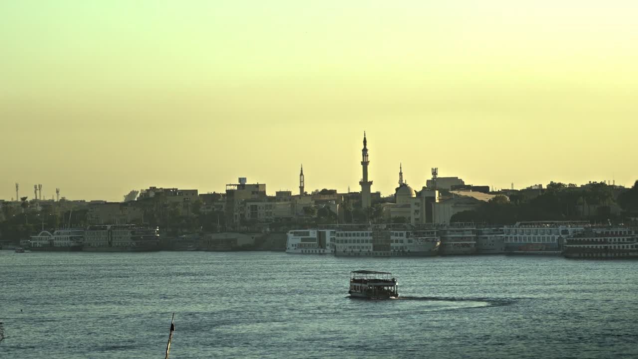 Il fiume Nilo a Luxor durante l'ora d'oro, con il sole che tramonta sull'acqua tranquilla. Sullo sfondo, lo skyline della sponda orientale presenta le distinte silhouette di un minareto di moschea