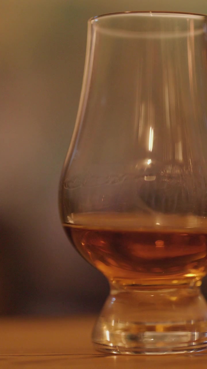 Una mano posa un bicchiere di whisky su un bancone di legno, con il liquido che si agita delicatamente. L'illuminazione calda e soffusa e la messa a fuoco ridotta creano un'atmosfera rilassata e invitante.