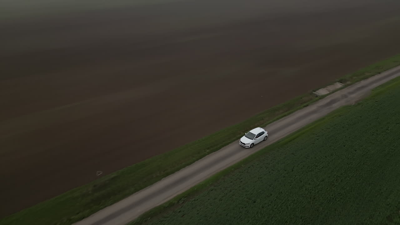 畑の間の田舎道を走る白い車、ドローンで追跡される広がる景色、悪天候の曇り空と霧
