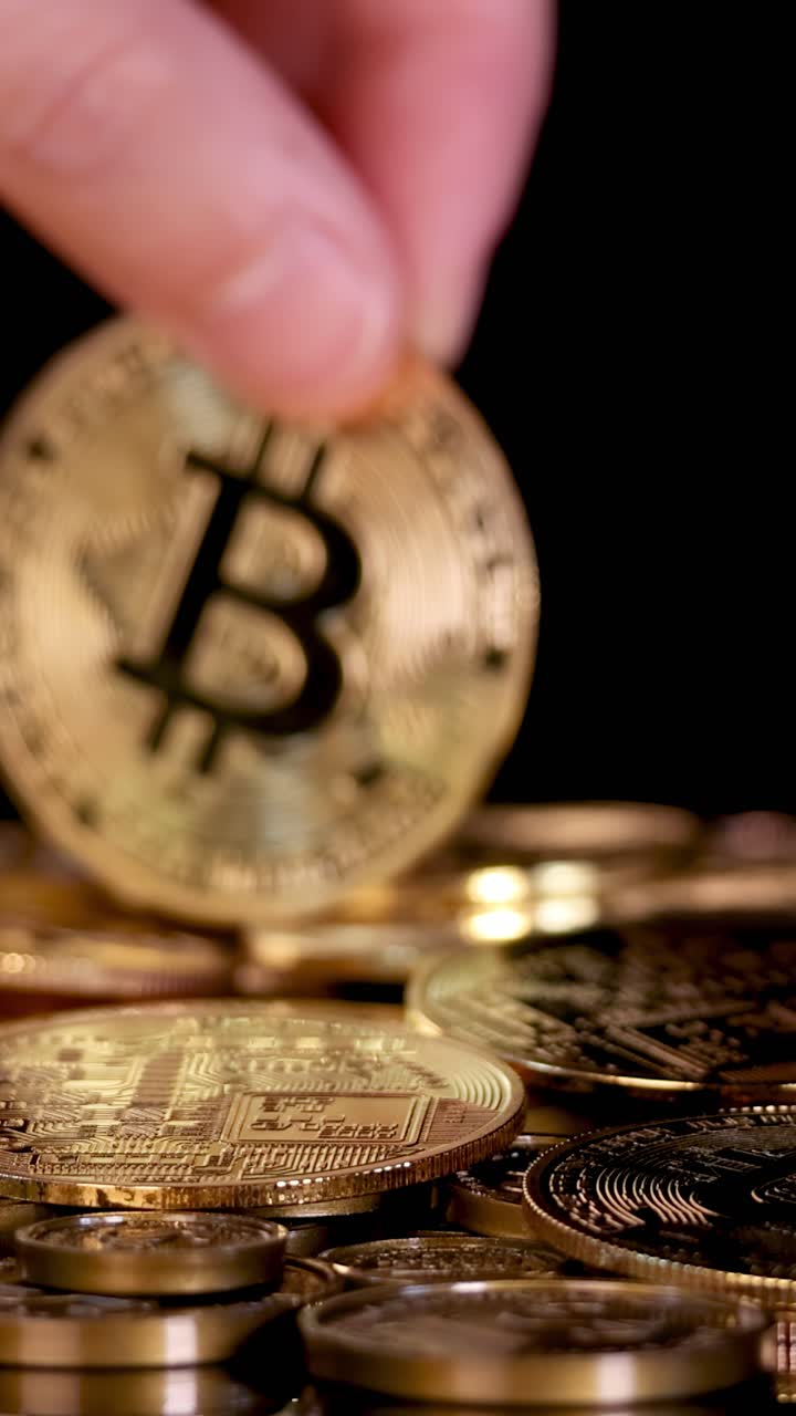 Mão em close-up deixa cair um token de bitcoin dourado sobre uma pilha de moedas, com iluminação dramática e fundo preto.