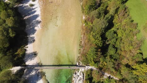 top view drone shot of tourist walking across rope bridge in alpine environment, clear river flows under bridge, green fields and trees on riverbeds, sunny summer day