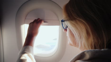 a woman opens the porthole curtain and looks out the window of the plane