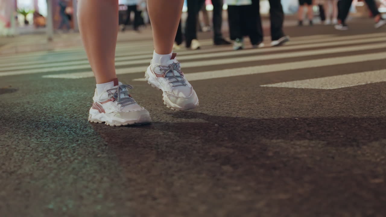 Night city crosswalk legs walking sneakers, closeup of casual footwear on textured asphalt under neon lights, pedestrian pausing between signals, blurred crowd and storefronts in background, evening