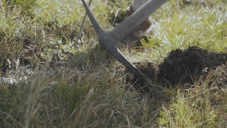 person digging on land grass land with help of axe, other person wears lather brown shoes