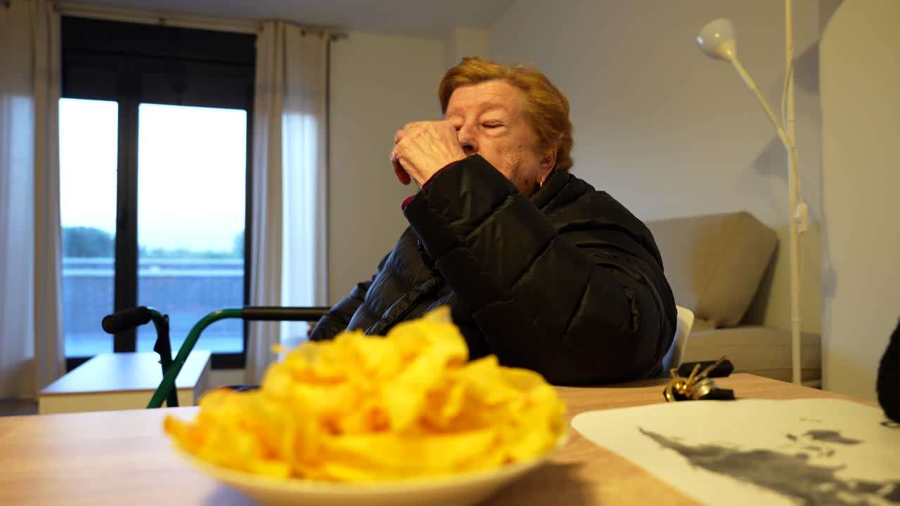 Granny and her young granddaughter snack on chips at living room table