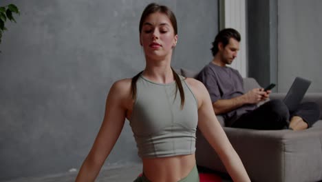 Close-up-a-confident-brunette-girl-in-a-gray-sports-top-and-green-pants-does-stretches-and-splits-on-a-red-sports-mat-Near-her-boyfriend-who-is-working-on-a-gray-laptop-Sitting-on-the-sofa-in-a-modern-apartment-at-home