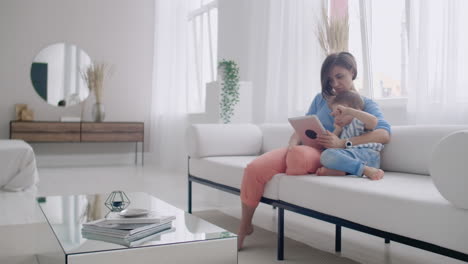 madre e hijo usando tableta mirando la pantalla y hablando en el sofá en una casa acogedora