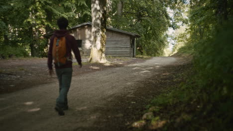 A-young-hiker-is-walking-on-forest-path-towards-the-forest-cabin,-admireing-tree-tops