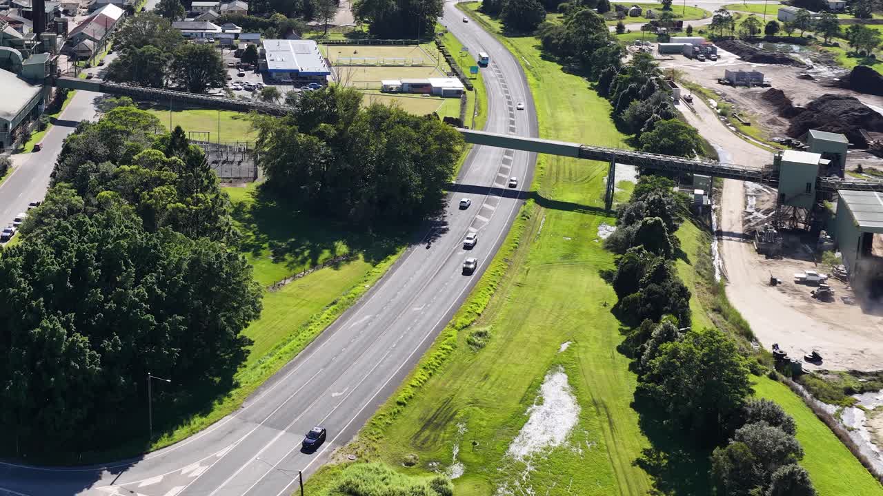 Aerial footage captures vehicles on a winding road surrounded by lush greenery and industrial structures under bright daylight