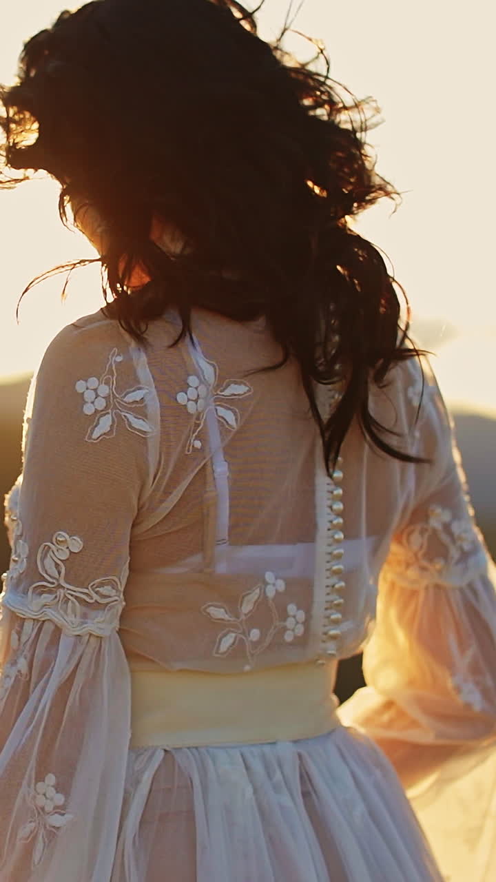 Long-haired lady wearing white dress standing high in the mountains. Lady is turning around at the backdrop of shining bright setting sun. Vertical video