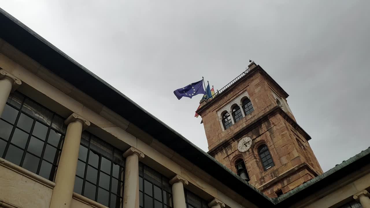 Clock tower on Oviedo's university building with waving flags on a cloudy day, seen in Oviedo's city center