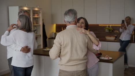 dois casais idosos felizes dançando na cozinha, enquanto no fundo desfocado uma mulher muito idosa os filma no celular 1