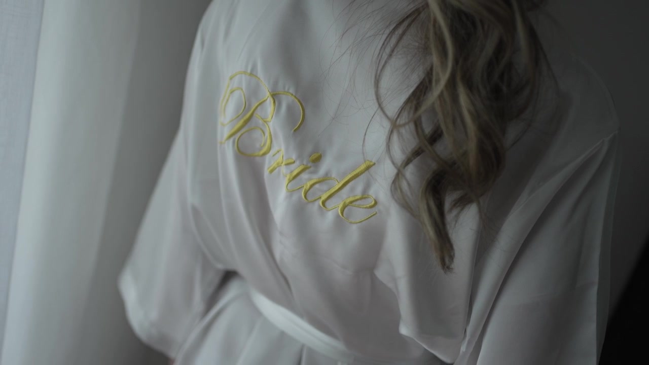 Close-up of the embroidered "Bride" lettering in gold on a white bridal robe, with soft hair draping over the shoulder