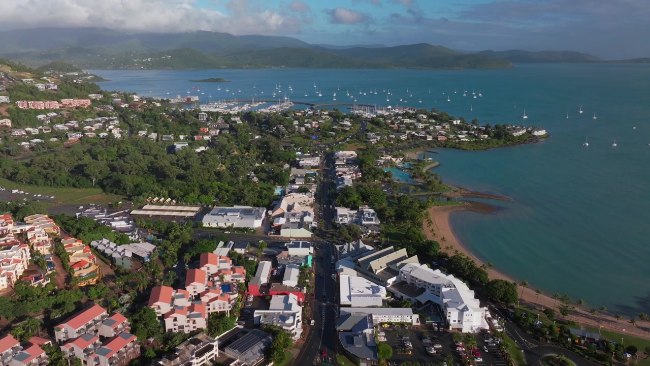 Pemandangan Drone Udara Laguna Teluk Pantai Airlie, Jantung Great Barrier Reef Whitsundays Whitehaven, menampilkan jalan utama, mobil, bangunan, matahari terbit, kabut pagi, Laut Koral yang cerah, marina, dermaga, dan perahu layar, bergerak maju ke atas.
