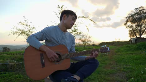 hombre asiático sentado con la pierna cruzada tocando la guitarra al aire libre al atardecer, vietnam