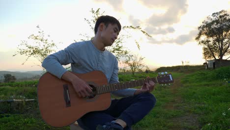 asian man sitting with leg crossed playing guitar at sunset outdoors, vietnam