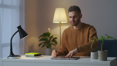 Trabajando-En-Casa,-Un-Joven-Está-Abriendo-Una-Computadora-Portátil-Y-Encendiendo-Una-Lámpara-De-Mesa-Por-La-Noche,-Trabajo-Remoto-En-Línea-Y-Navegación-Independiente-Por-Internet