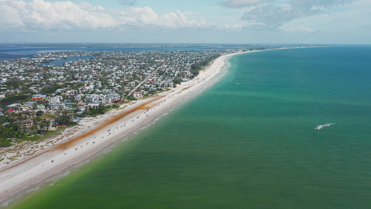 Turquoise waters stretch along the sandy shoreline of Anna Maria Island, with residential neighborhoods filling the coast under a bright summer sky.
