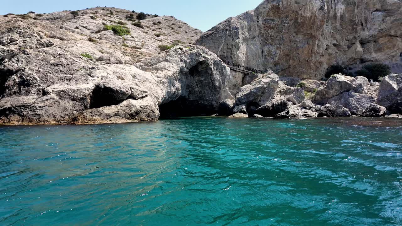 A boat travels along the coast of Crimea, passing by rocky cliffs and a cave, on a bright, sunny day