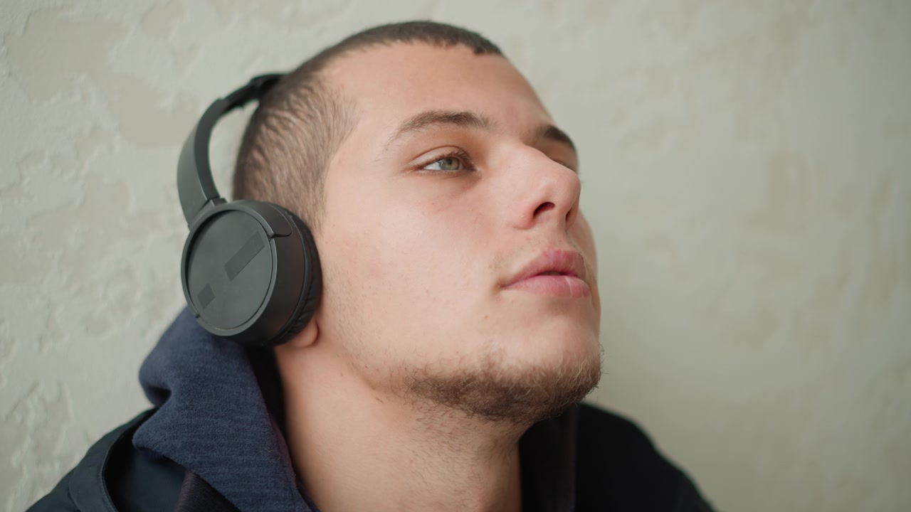 Primo piano di un uomo che indossa cuffie nere over-ear e abiti scuri contro una parete texturizzata di colore chiaro. Il soggetto rimane immobile con movimenti minimi, mantenendo la stessa posa. Illuminazione soffusa.
