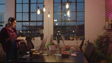 young caucasian business woman student working late using computer typing research project browsing online writing notes brainstorming deadline in trendy office at night wearing headphones