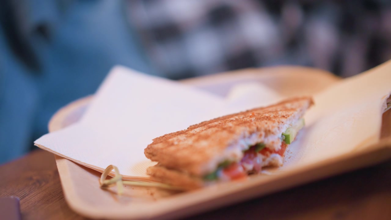 Primo piano di un toast con ripieno visibile di pomodoro e verdure verdi su carta da forno, posizionato diagonalmente su un vassoio di legno. Lo sfondo presenta elementi sfocati, incluso il blu.