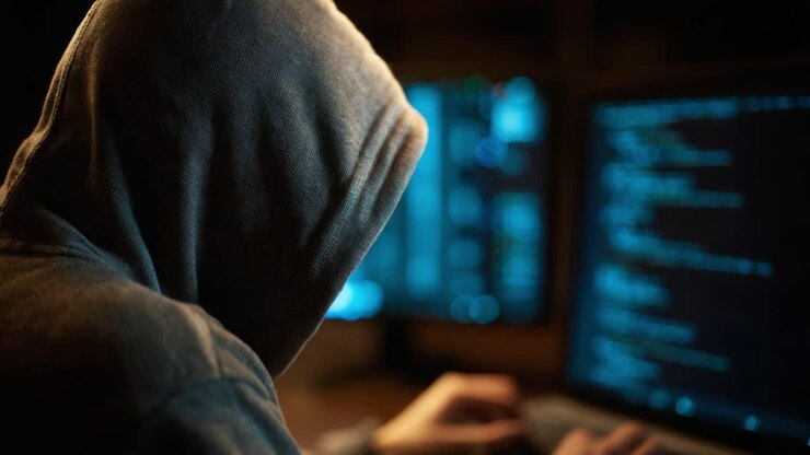 Una figura misteriosa con una felpa con cappuccio impegnata in un intenso lavoro al computer, circondata da monitor oscurati che mostrano righe di codice e analisi di dati digitali