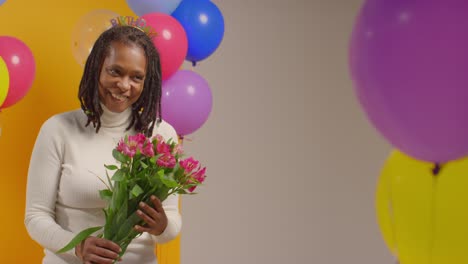 studio portrait of woman wearing birthday headband holding bunch of flowers celebrating with balloons 3