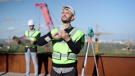 disparo medio de un hombre caucásico con sombrero duro mirando hacia arriba al sol esparciendo dinero sonriendo. feliz ingeniero rico confiado en el sitio de construcción al aire libre con compañero de trabajo borroso en el fondo.