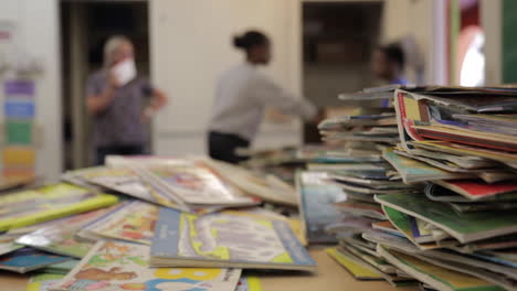 Teachers-Talk-in-Empty-Classroom-in-Distance,-Pile-of-Books-on-Desk-in-Foreground,-Depth