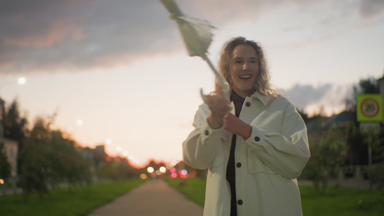 Cheerful woman in white coat playfully swinging folded umbrella along paved path at sunset with vibrant bokeh lights, residential buildings, parked cars, and greenery