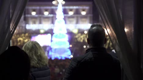 tiro panorámico manual en cámara lenta de personas mirando por una ventana abierta con cortinas en un festival de navidad en medina sidonia en cádiz españa en la plaza un árbol de navidad que brilla intensamente en azul