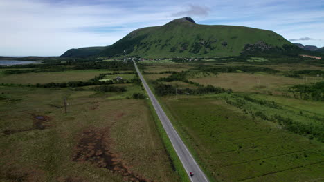 Aerial-dolly-forward-view-of-a-mountain-with-a-small-town-at-the-base-near-the-water-and-a-straight-country-highway-with-a-single-car-on-it-in-Northern-Norway-near-Lofoten-Norway