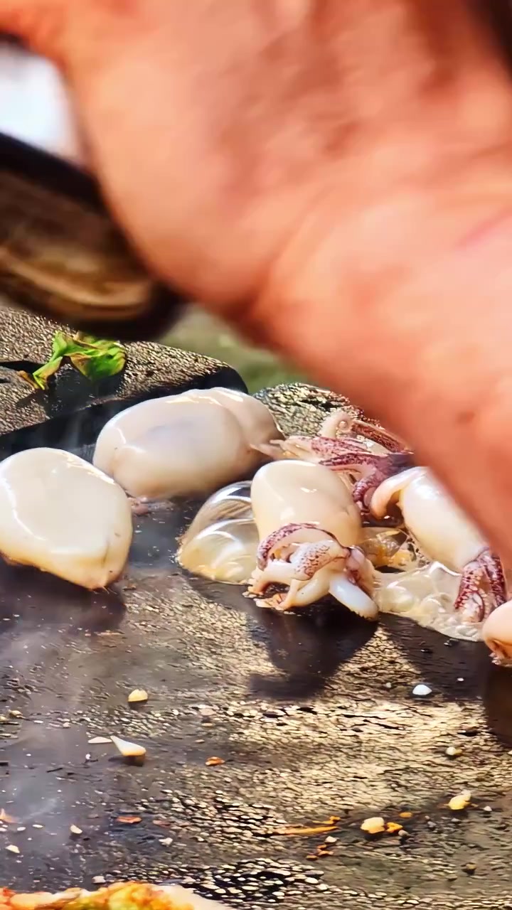 Close up of a hand seasoning baby squid and mussels with garlic on a hot black griddle alongside herb and meat flatbreads cooking at an outdoor street food festival stall in Riga Latvia