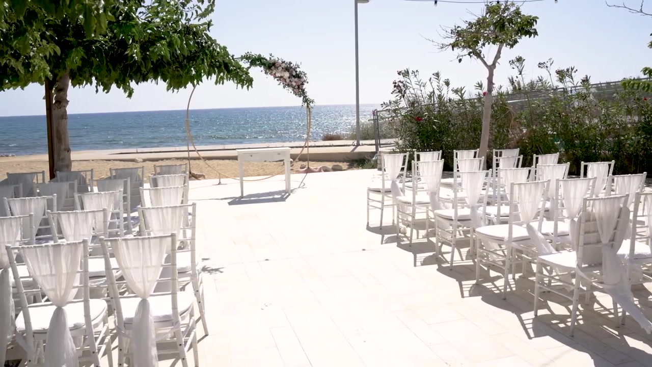Una splendida ambientazione per una cerimonia di matrimonio si svolge in una giornata di sole in spiaggia. Sedie bianche allineate di fronte all'oceano, incorniciate dalla vegetazione e da un cielo azzurro limpido.
