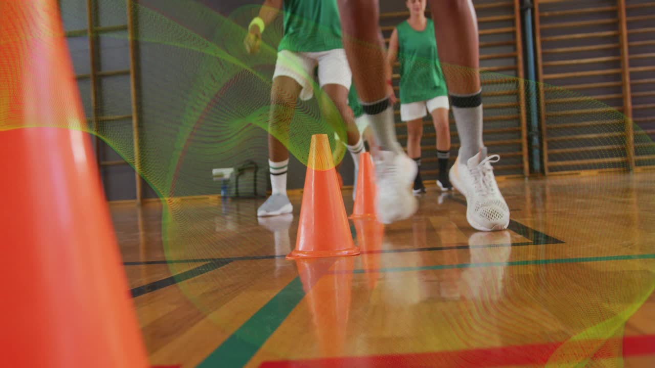 Atleta líder pegando a bola de basquete, driblando entre cones, com a equipe seguindo em treino de revezamento para agilidade.