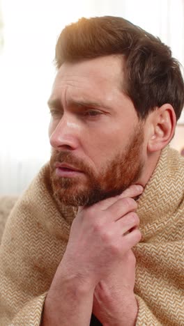 young man sitting on the sofa at home wrapped in a blanket coughing holding throat feeling pain