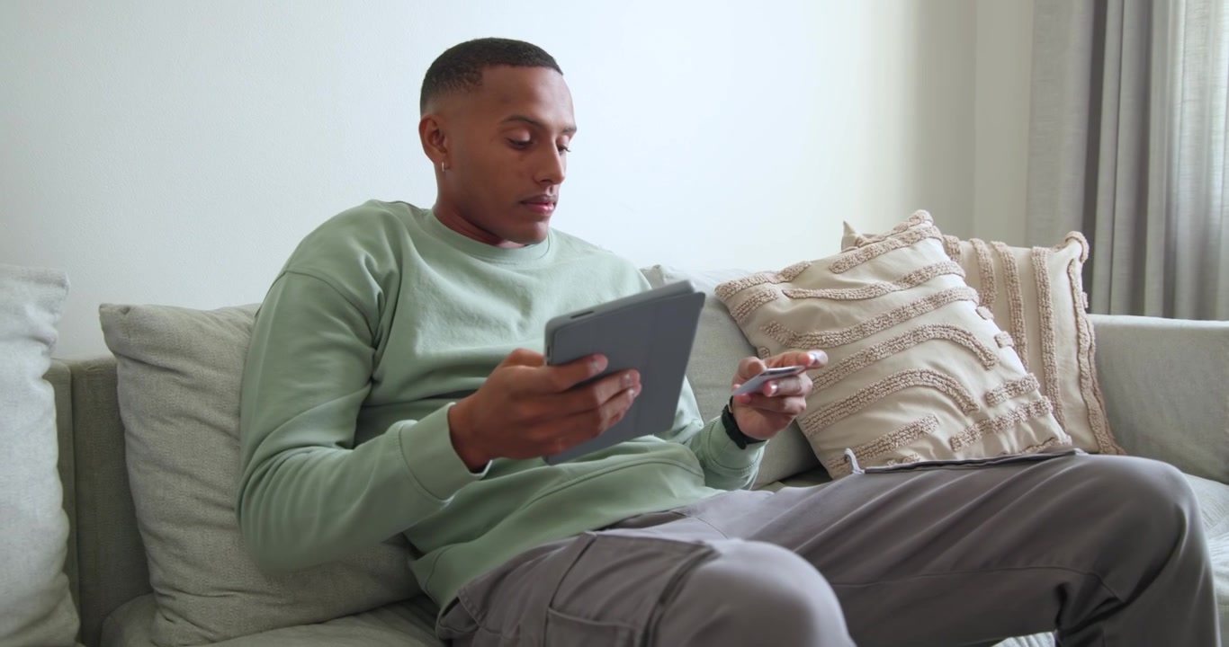 African American man leaning on sofa at home holding tablet and comparing small card with device