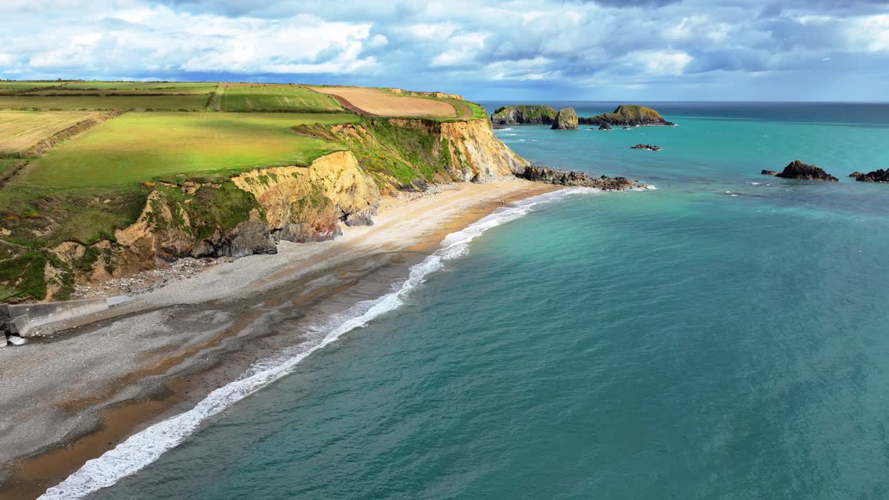 Drone establishing shot of cloud shadows drifting along beach with emerald green seas and gentle waves lapping on shingle beach Copper Coast in Waterford Ireland