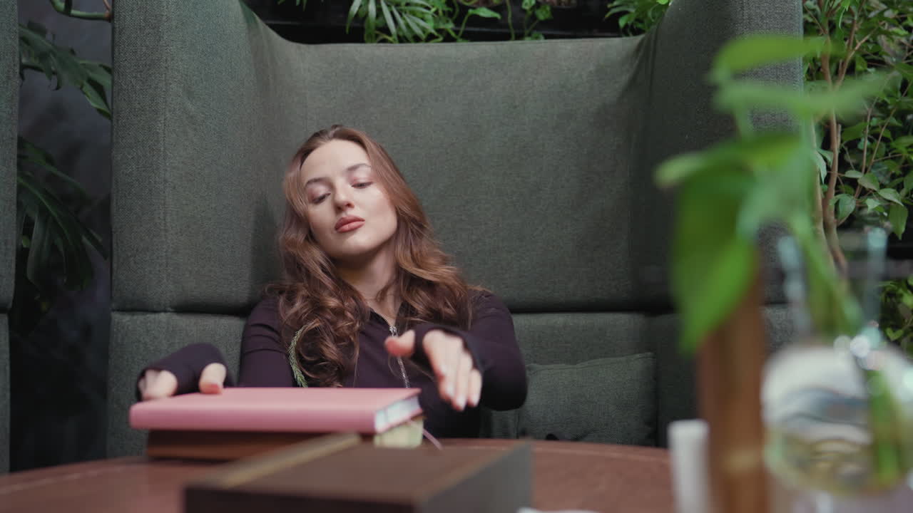 Blonde woman with soft brown-blonde hair in black top sits in green booth placing two books and pen on table while opening pink notebook, surrounded by indoor plants and calm cozy atmosphere