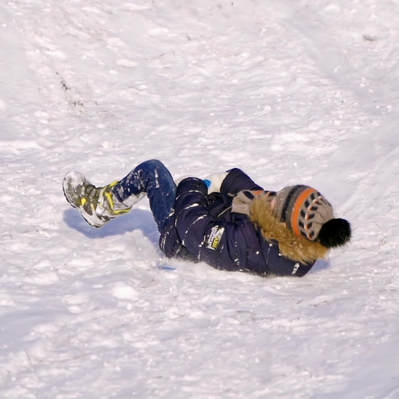 Cheerful kid on a snowy slide. Little boy on plastic sled riding down from white hill in winter. Cute child having fun at winter holidays. Slow motion.