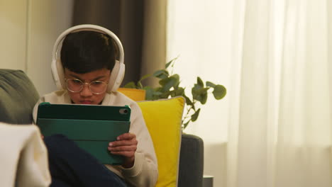 young boy sitting on sofa at home wearing wireless headphones playing games or streaming onto digital tablet