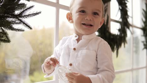 Adorable-Niño-Rubio-Con-Camisa-Blanca-Parado-En-El-Alféizar-De-La-Ventana-Sosteniendo-Un-Juguete-De-Navidad-Y-Sonriendo.-La-Ventana-Está-Decorada