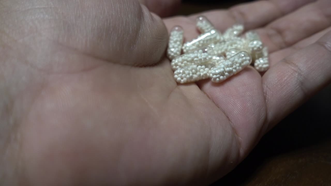 primer plano de una mano mientras se vierte en ellas varias cápsulas medicinales transparentes con pellets blancos en su interior