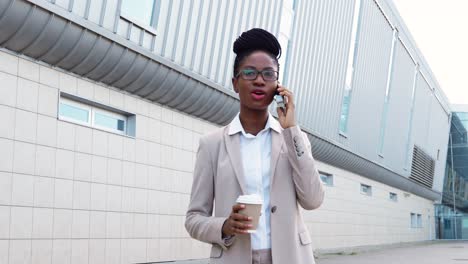 african american businesswoman in stylish clothes and glasses talking on the smartphone and holding coffee to go in the street