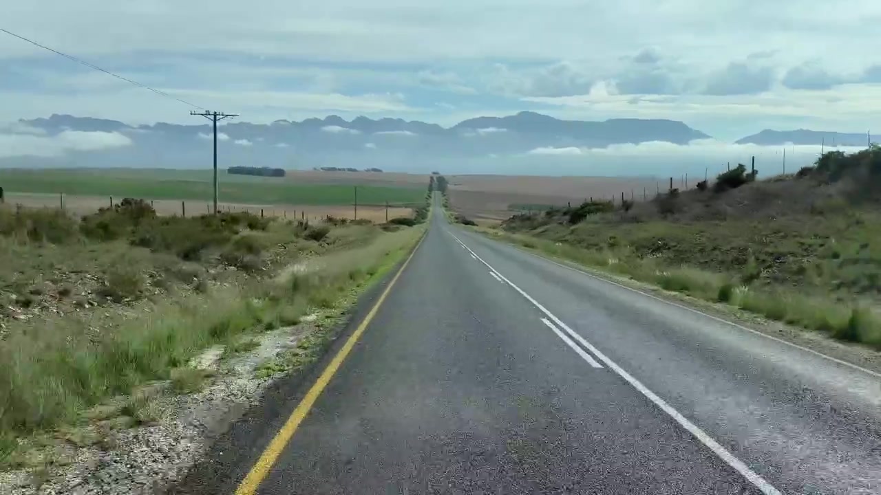 Estrada longa que se estende pela paisagem rural sul-africana com um cenário de montanhas.