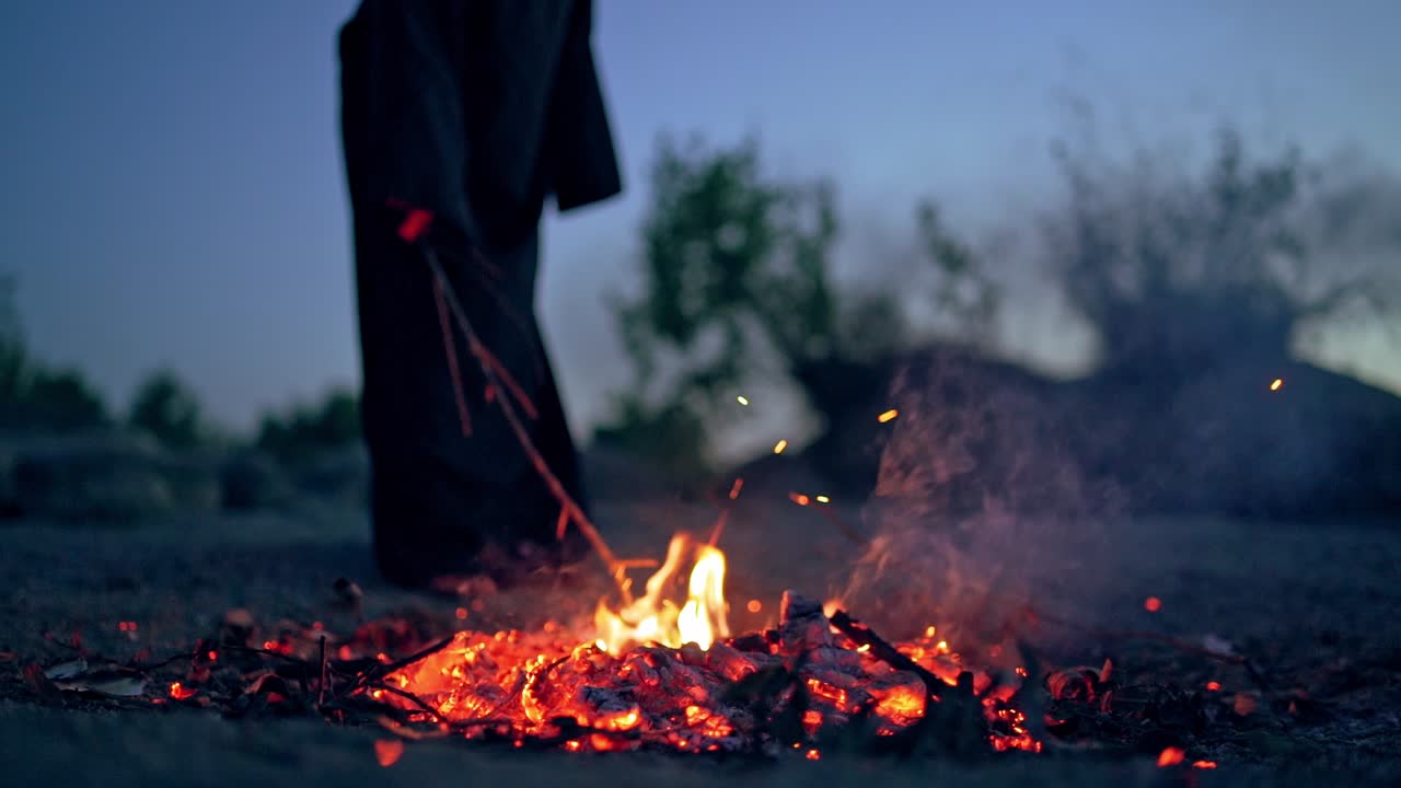 Scary witch evil with a stick near the fire. Dark death next to smoldered logs of fire in the evening. Horror atmosphere outdoors.