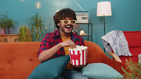 Indian-man-sits-on-sofa-eating-popcorn-and-watching-interesting-TV-serial,-sport-game-online-at-home